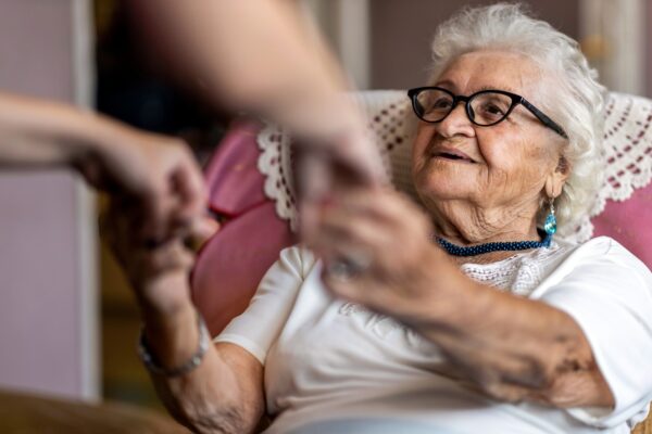 Female home carer supporting old woman to stand up from the armchair